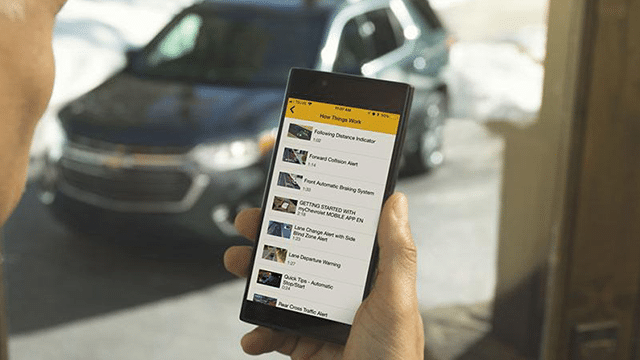 Driver holding a phone showing the GM mobile app, with a vehicle in the driveway behind