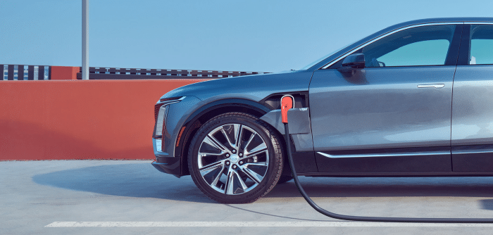 Dark grey Cadillac electric SUV plugged in at a public charging spot beside an orange wall