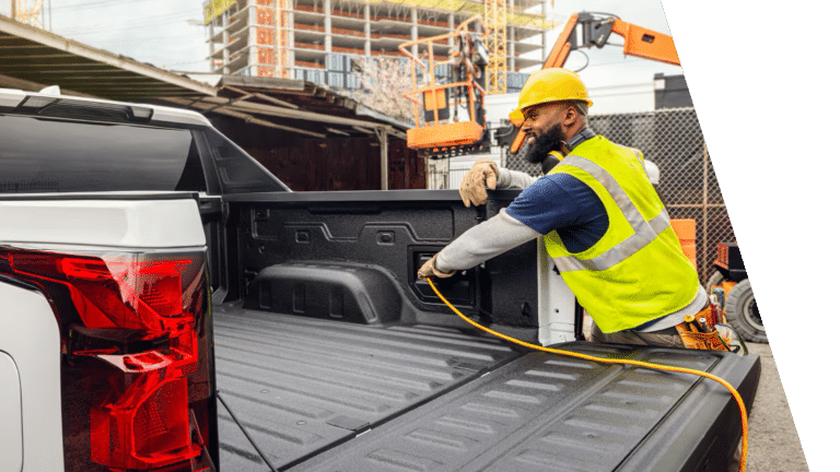 Worker in a hard hat and safety vest plugging a power cord into the bed outlet of an electric pickup truck on a job site