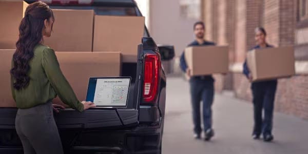 Woman working on a laptop on a pickup truck tailgate while two delivery workers carry boxes toward the truck