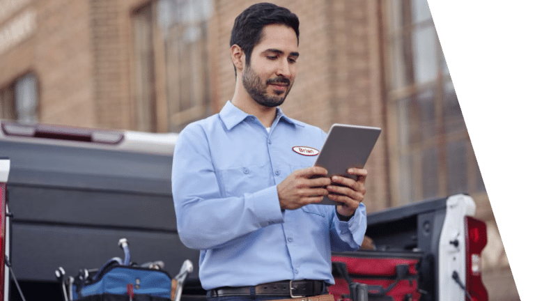 Service technician in a light blue work shirt checking a tablet beside an open pickup truck tailgate loaded with tool bags