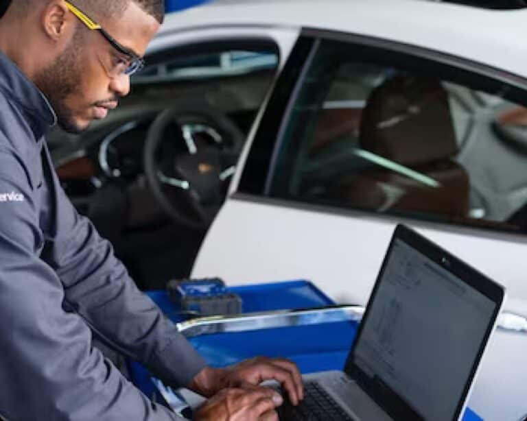 Certified Service technician running a diagnostic scan on a laptop beside a white Chevrolet sedan