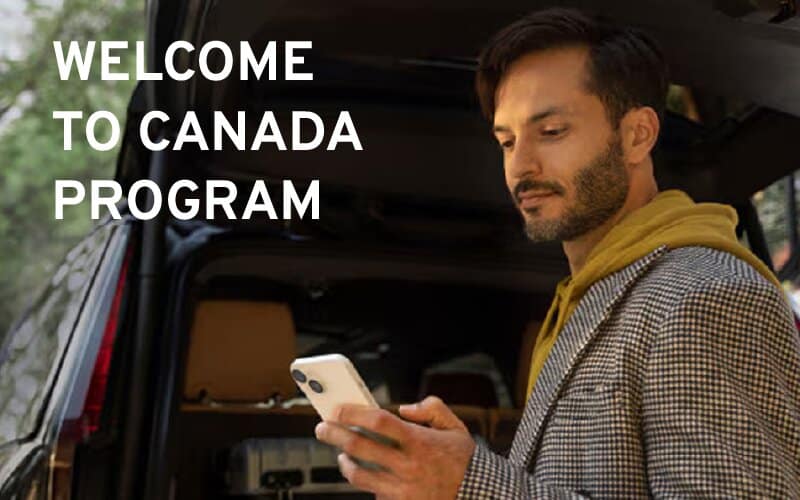 Man checking his phone beside the open tailgate of a vehicle packed with luggage, under the words Welcome to Canada Program