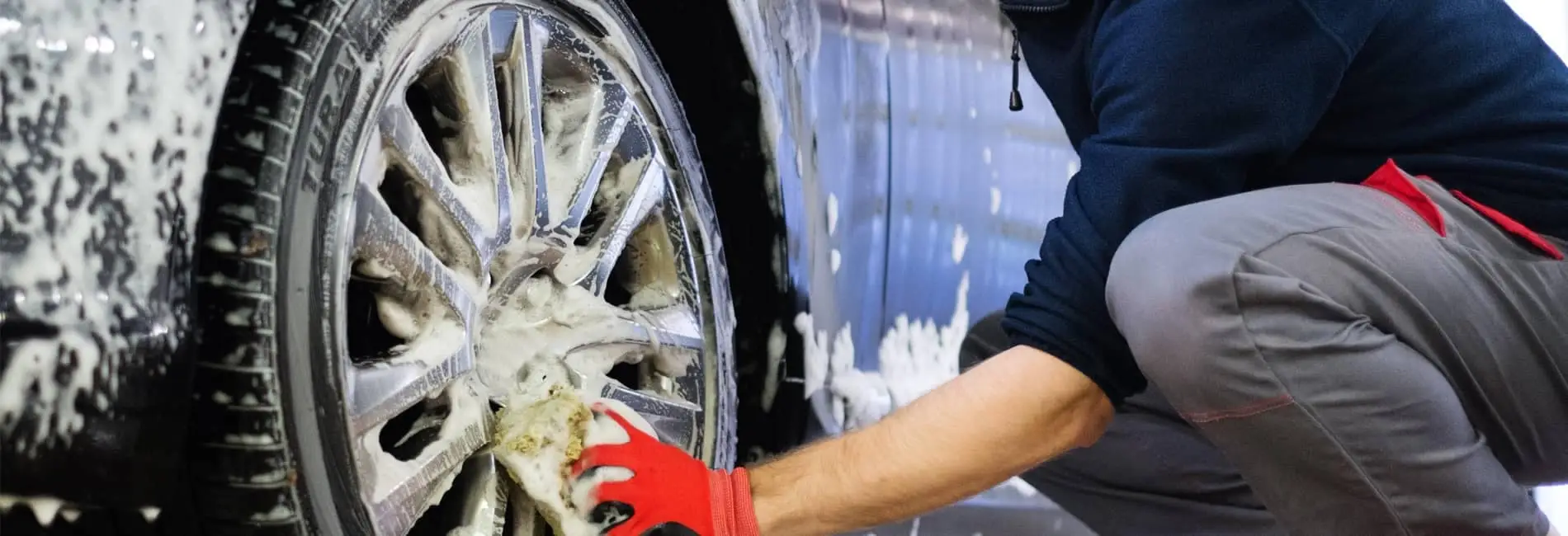 Person in work clothes crouching to wash a vehicle alloy wheel with a soapy sponge