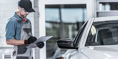 Service technician with a clipboard inspecting a white vehicle in a service bay