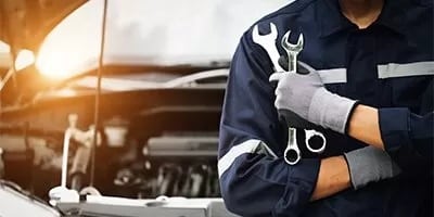 Technician in a dark blue uniform holding two wrenches in front of an open car hood